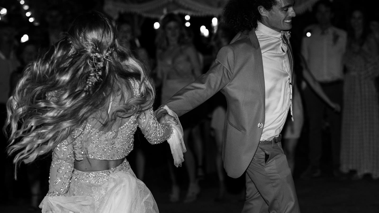 A bride and groom dancing together as guests look on, string lights overhead